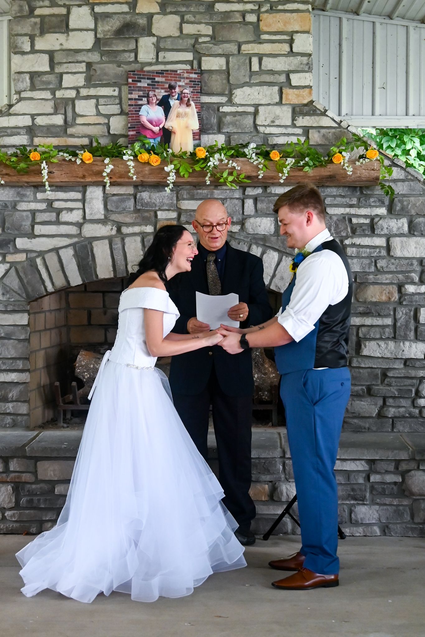 Bride and groom exchange vows before an officiant at a stone fireplace altar decorated with greenery and flowers.
