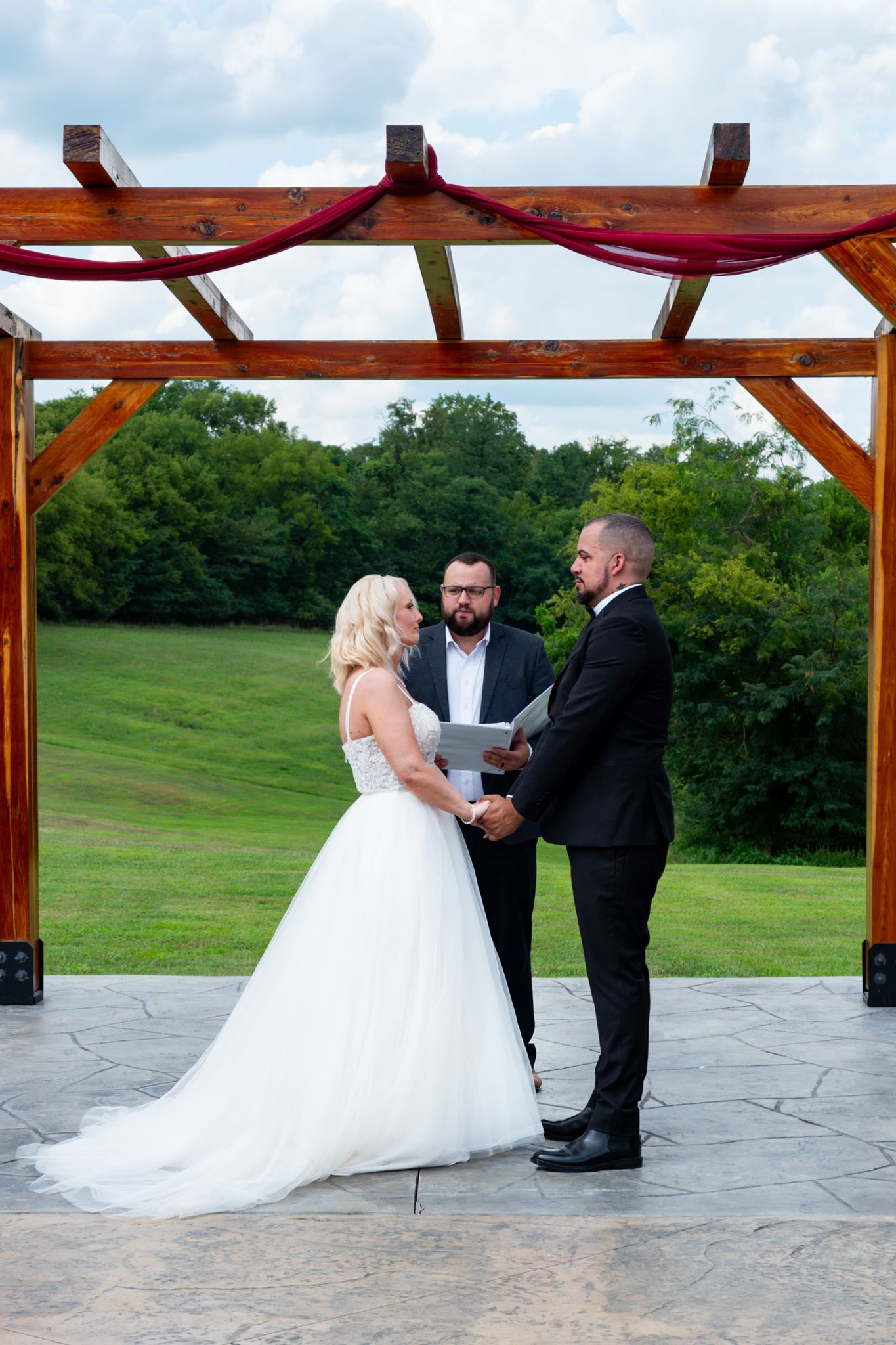 Bride and groom exchange vows under a wooden pergola with red draping, officiant holding book, green hills backdrop.