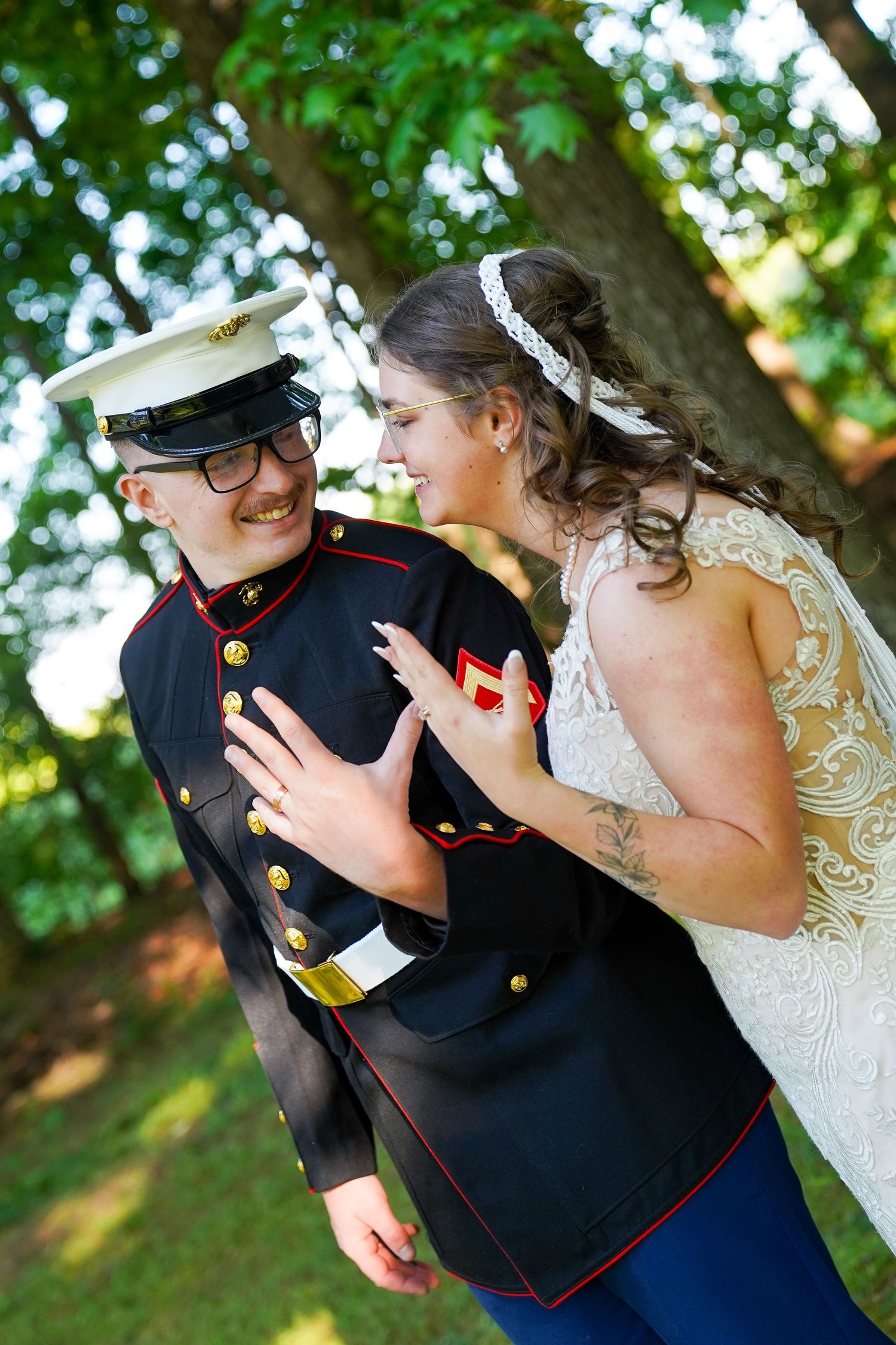 Bride in lace wedding dress laughing with groom in Marine dress blues uniform outdoors under green trees.