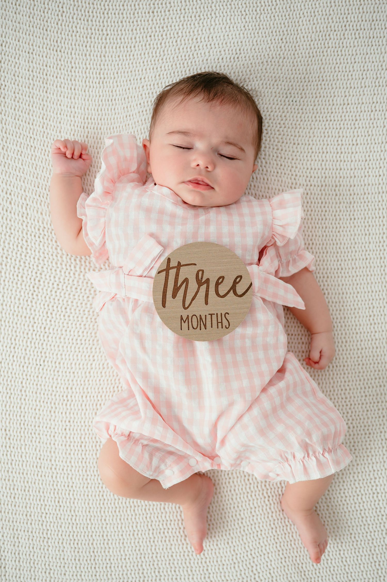 A sleeping 3-month-old baby girl in a pink gingham dress lies on a white blanket with a wooden milestone disc.