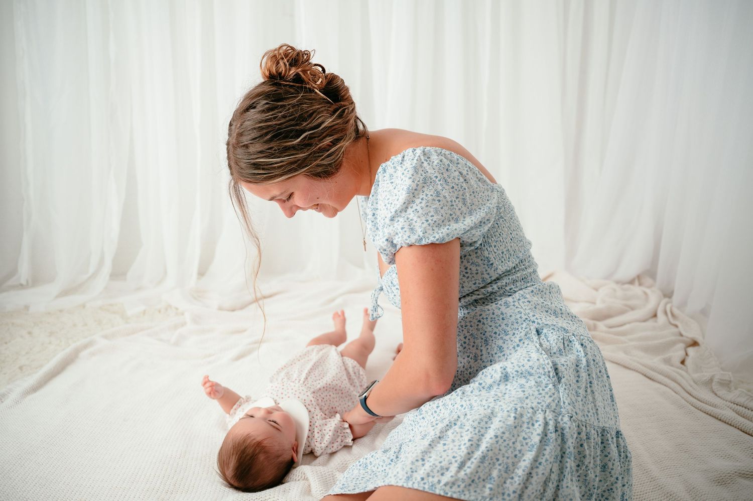 Mother in floral dress tenderly interacting with newborn baby lying on white bed during professional photoshoot.