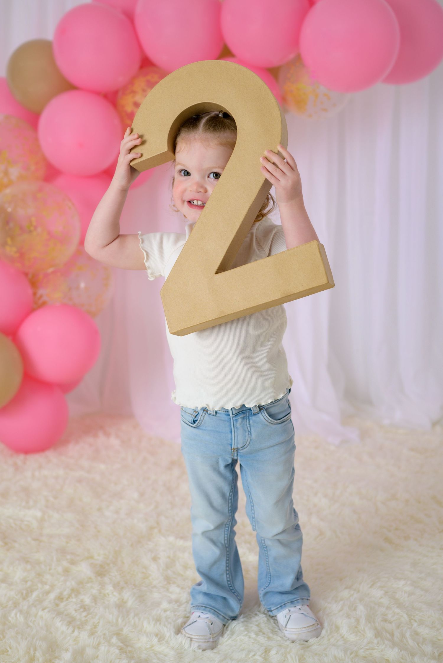 Cute toddler girl peeking around oversized gold number 2 prop with pink balloon backdrop.