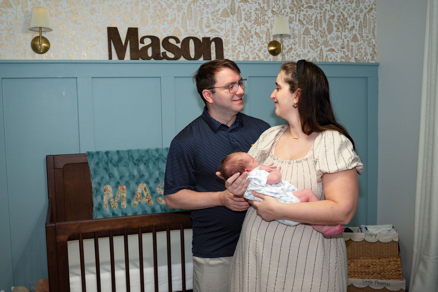 New parents smile lovingly at each other while holding their newborn baby in a nursery decorated with the name Mason on the wall.