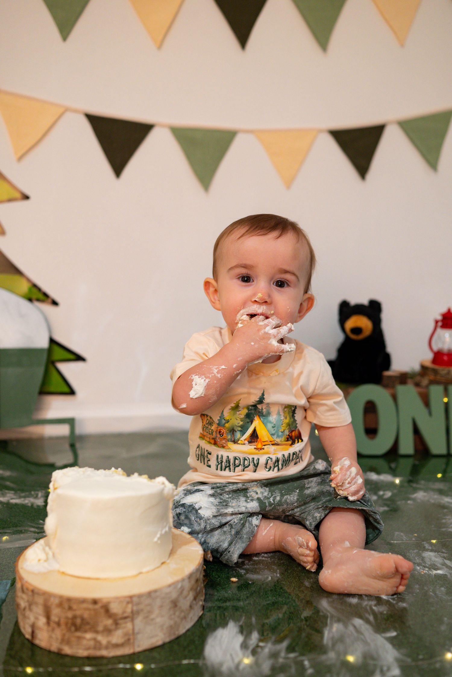 Baby boy with frosting on his face sits next to his white smash cake at a camping-themed first birthday.