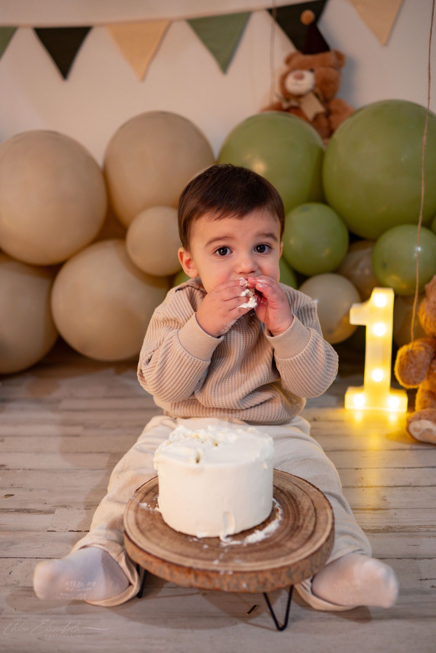 Baby boy celebrates first birthday smashing and eating white cake surrounded by green and beige balloons.