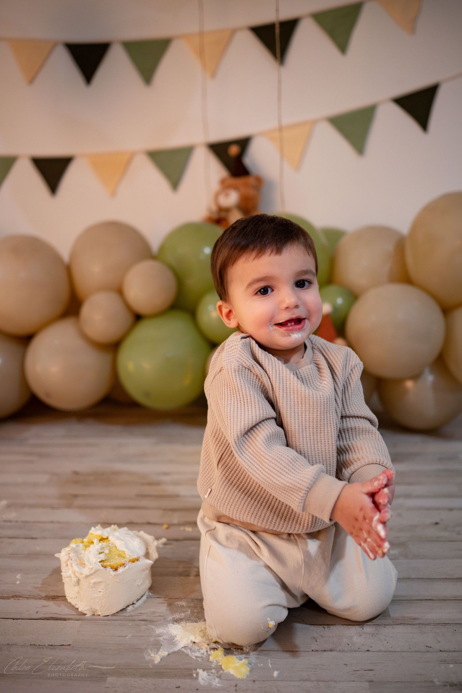 Happy toddler boy celebrating first birthday cake smash with balloons and bunting decorations.