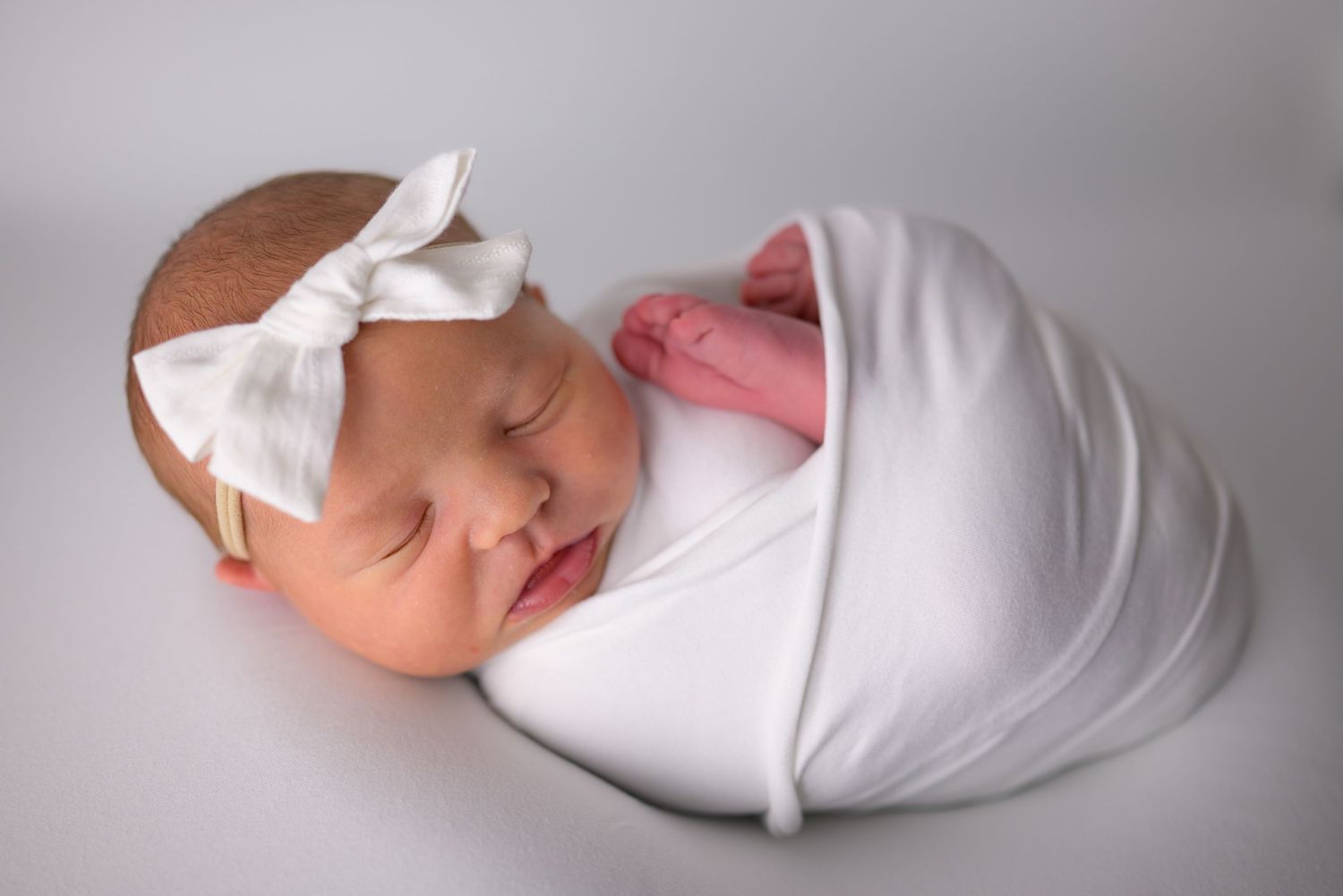 Newborn baby sleeping peacefully swaddled in white wrap with a white bow headband.