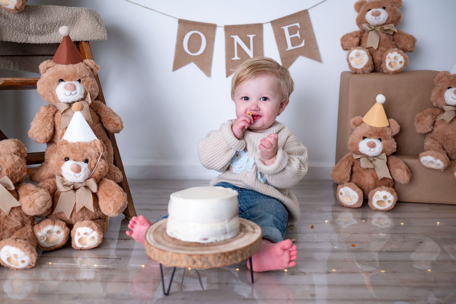 Smiling baby boy in beige sweater sits near white smash cake with teddy bear decorations and ONE banner.