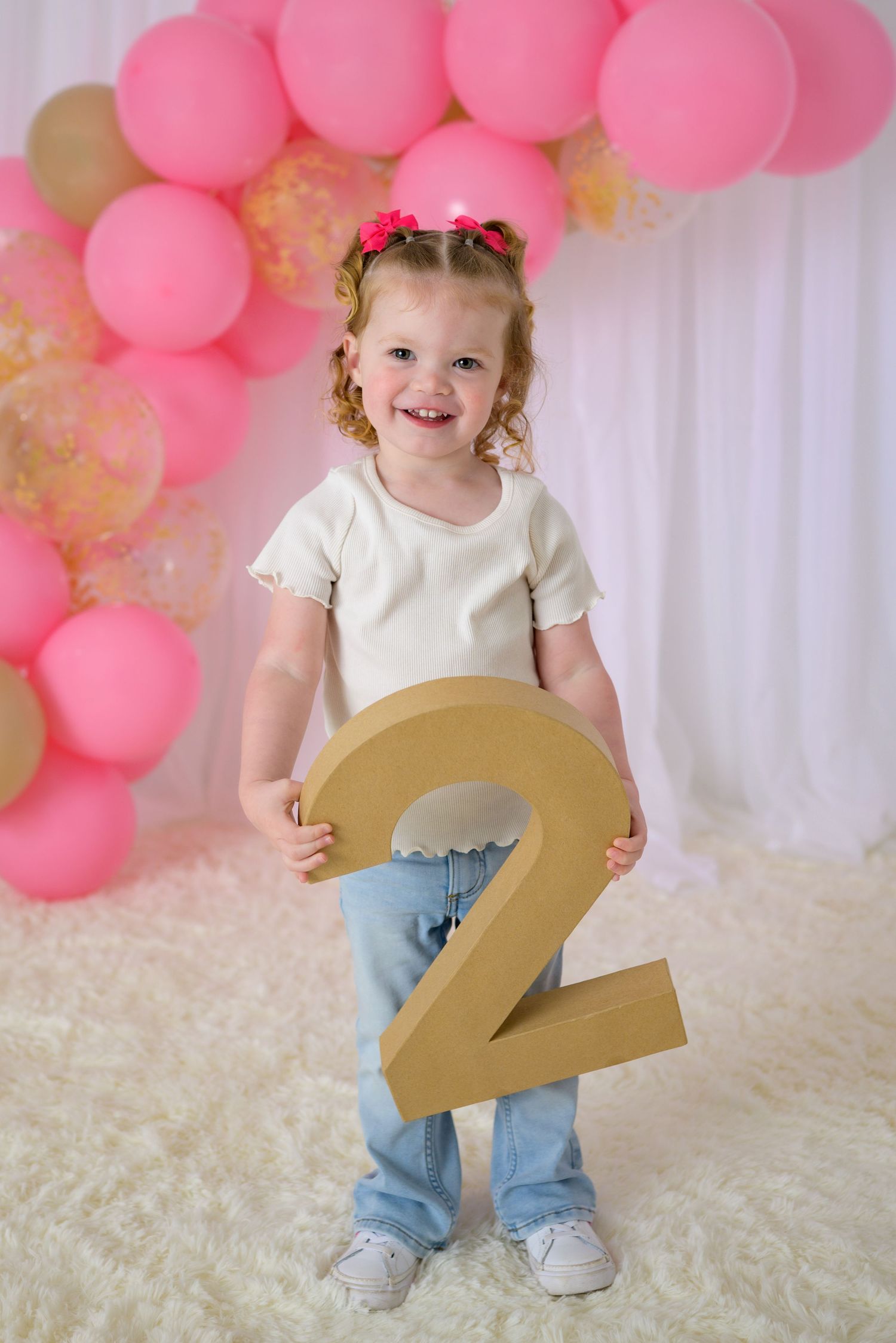 Happy toddler girl holding large gold number 2 in front of pink balloon arch for birthday photos.