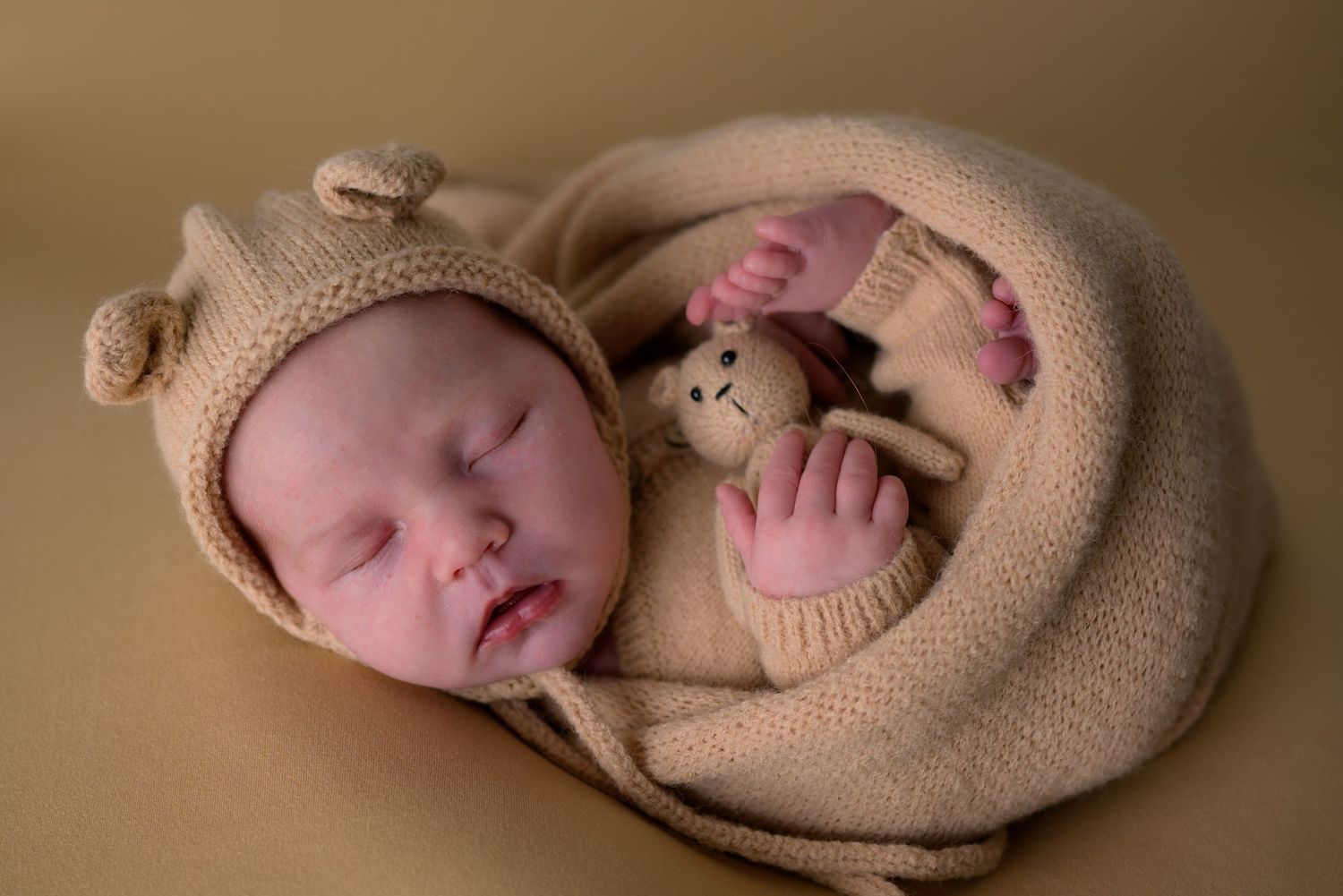 Sleeping newborn baby in beige knit wrap and bear bonnet holding a stuffed animal toy.