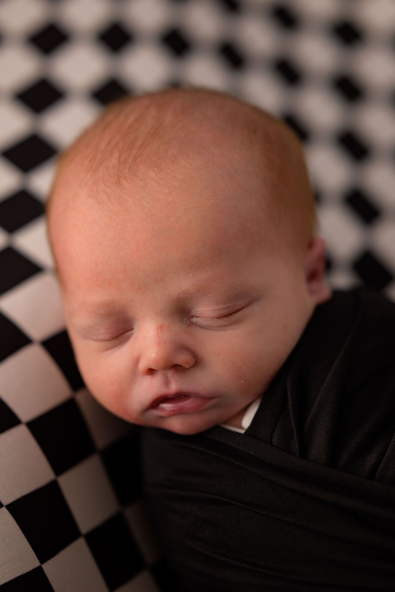 Sleeping newborn baby swaddled in black wrap on a black and white checkered background.