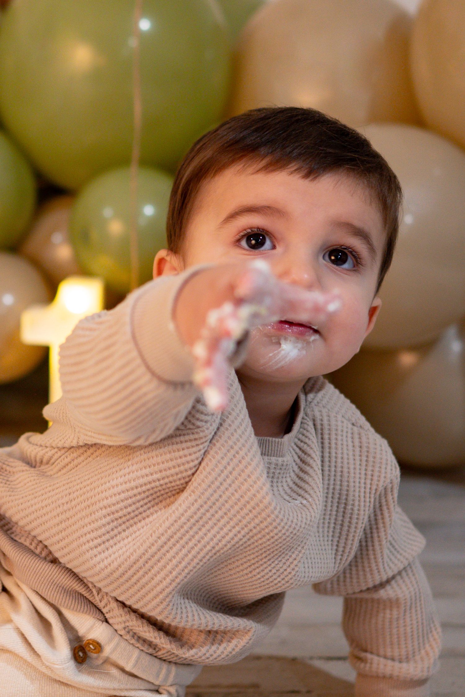 Curious one-year-old boy with cake on his face crawls toward camera with green and gold balloons behind him.