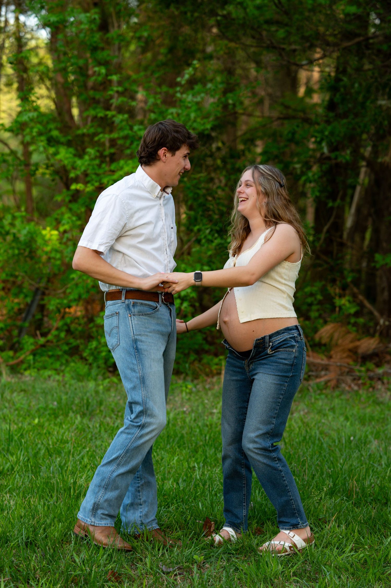 Smiling pregnant couple holding hands dancing together in lush green field. Venue: Cotton Wood Chapel