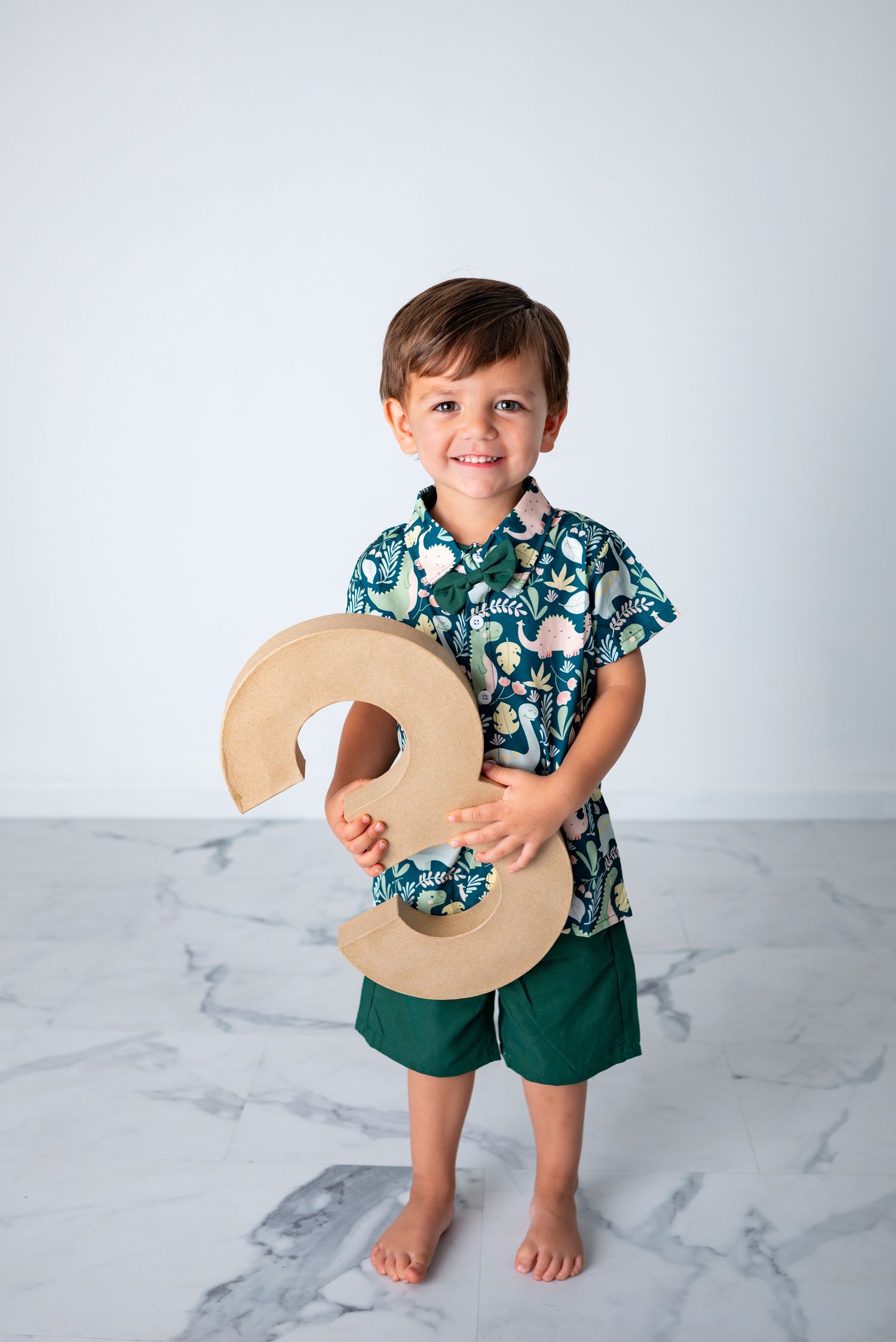 Smiling toddler boy holding a wooden number 3, wearing a green floral shirt and green shorts, standing on marble floor.