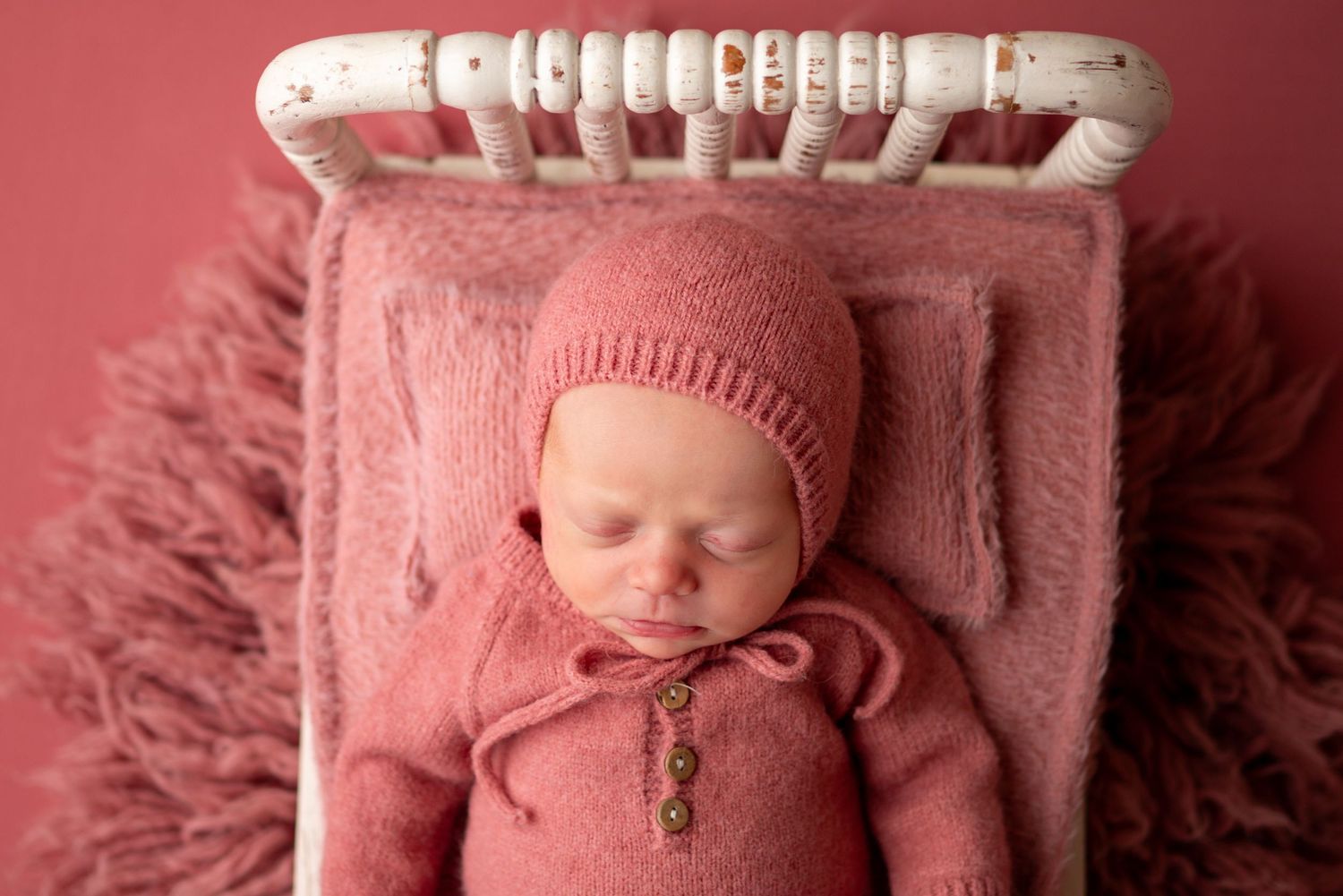 Sleeping newborn baby in pink knit romper and bonnet on a small white bed with pink backdrop.