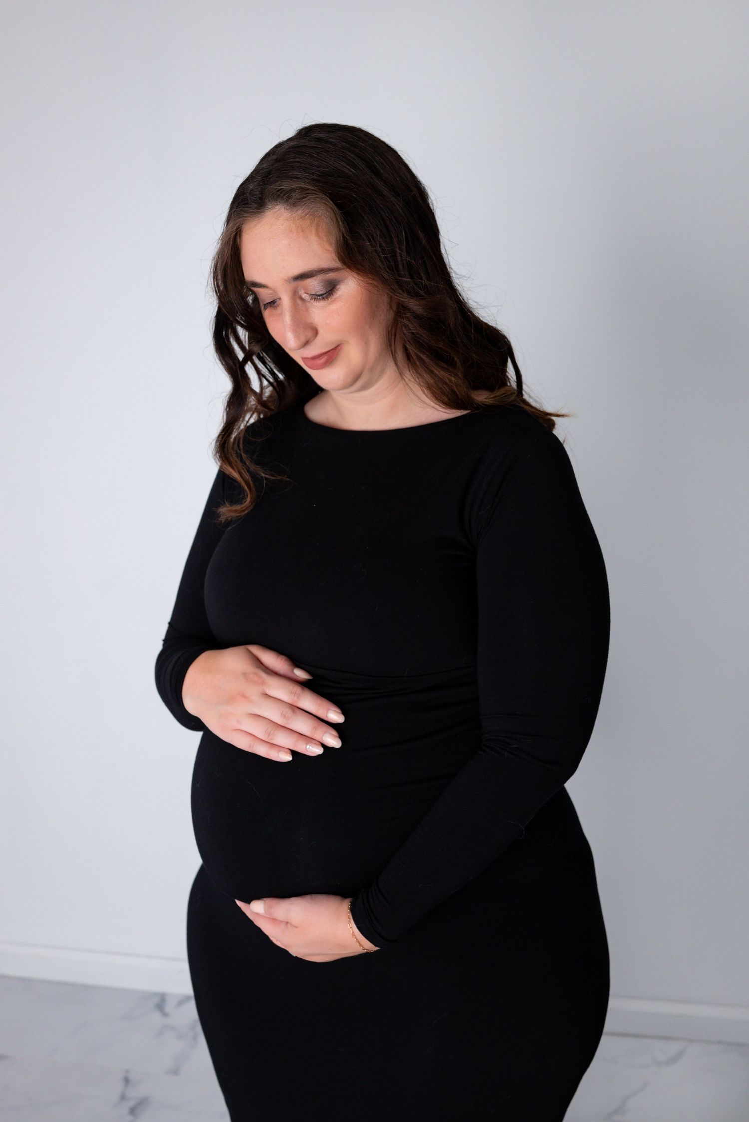 A pregnant woman in a black dress cradles her baby bump while looking down in a bright white studio.