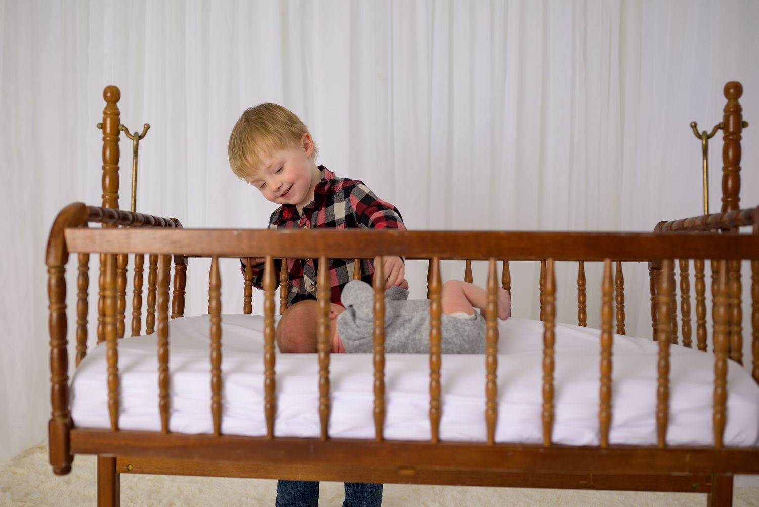 Smiling toddler boy in plaid shirt leaning over wooden crib railing looking at newborn sibling.