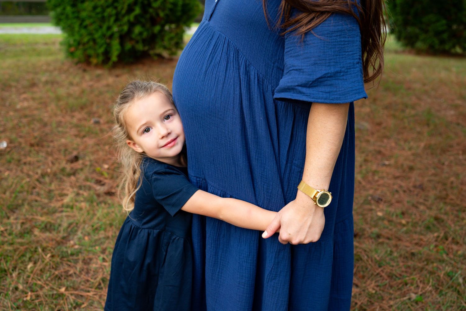 A young girl in a navy dress hugs her pregnant mother's baby bump during an outdoor maternity photo session.