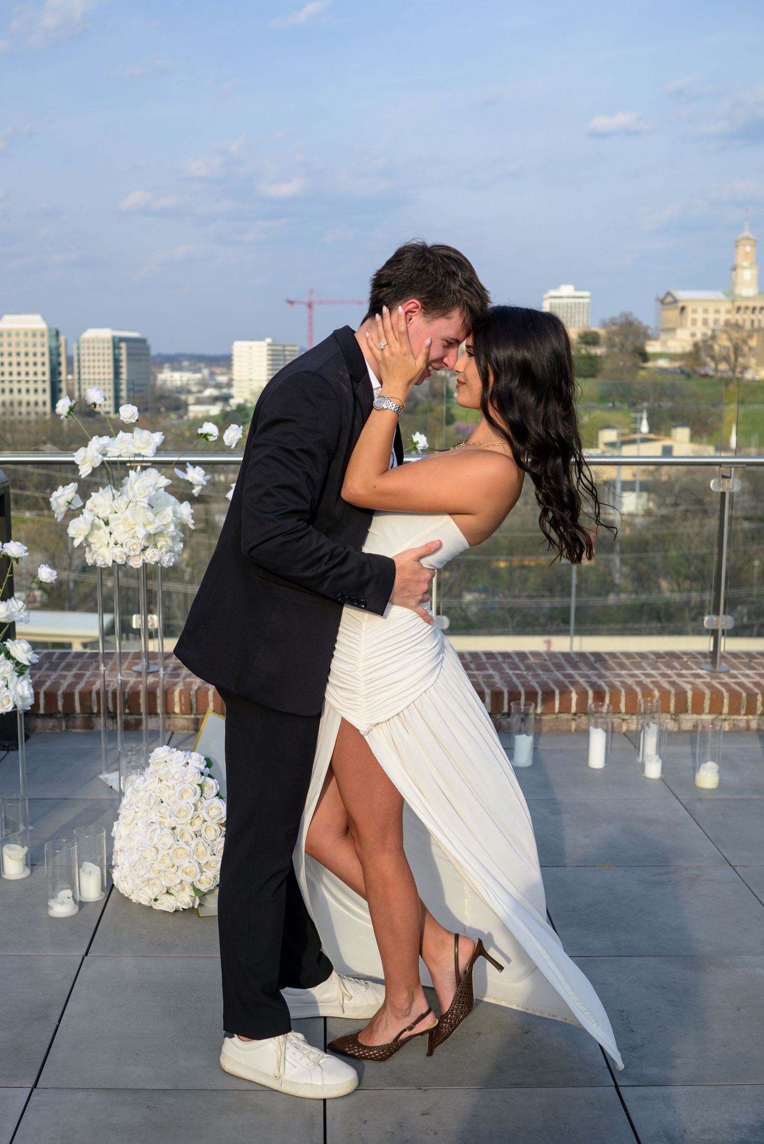 Engaged couple sharing intimate moment on rooftop venue with city skyline backdrop and floral decor.