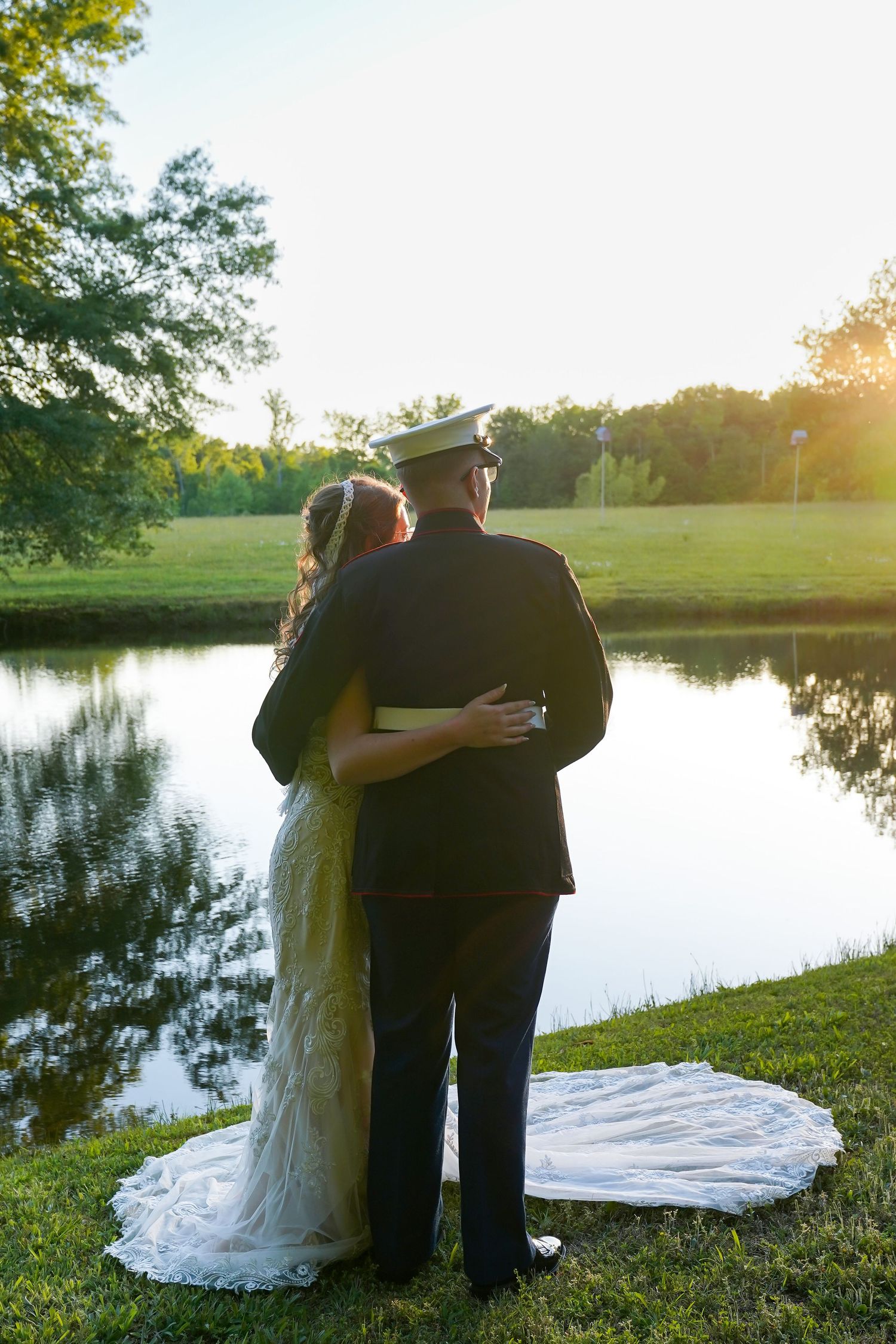 Bride and groom in military uniform embrace by a serene pond at sunset, with her white gown trailing on lush green grass.
