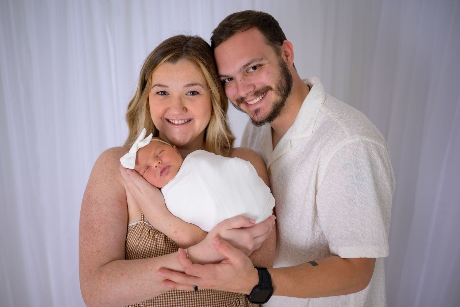 Happy couple smiles while cradling their swaddled newborn baby wrapped in white against a white backdrop.