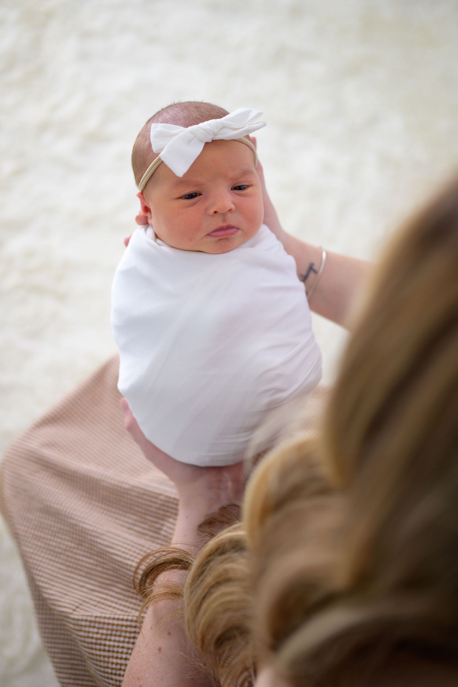 Alert newborn baby girl in white swaddle and bow headband being held up by her mother.