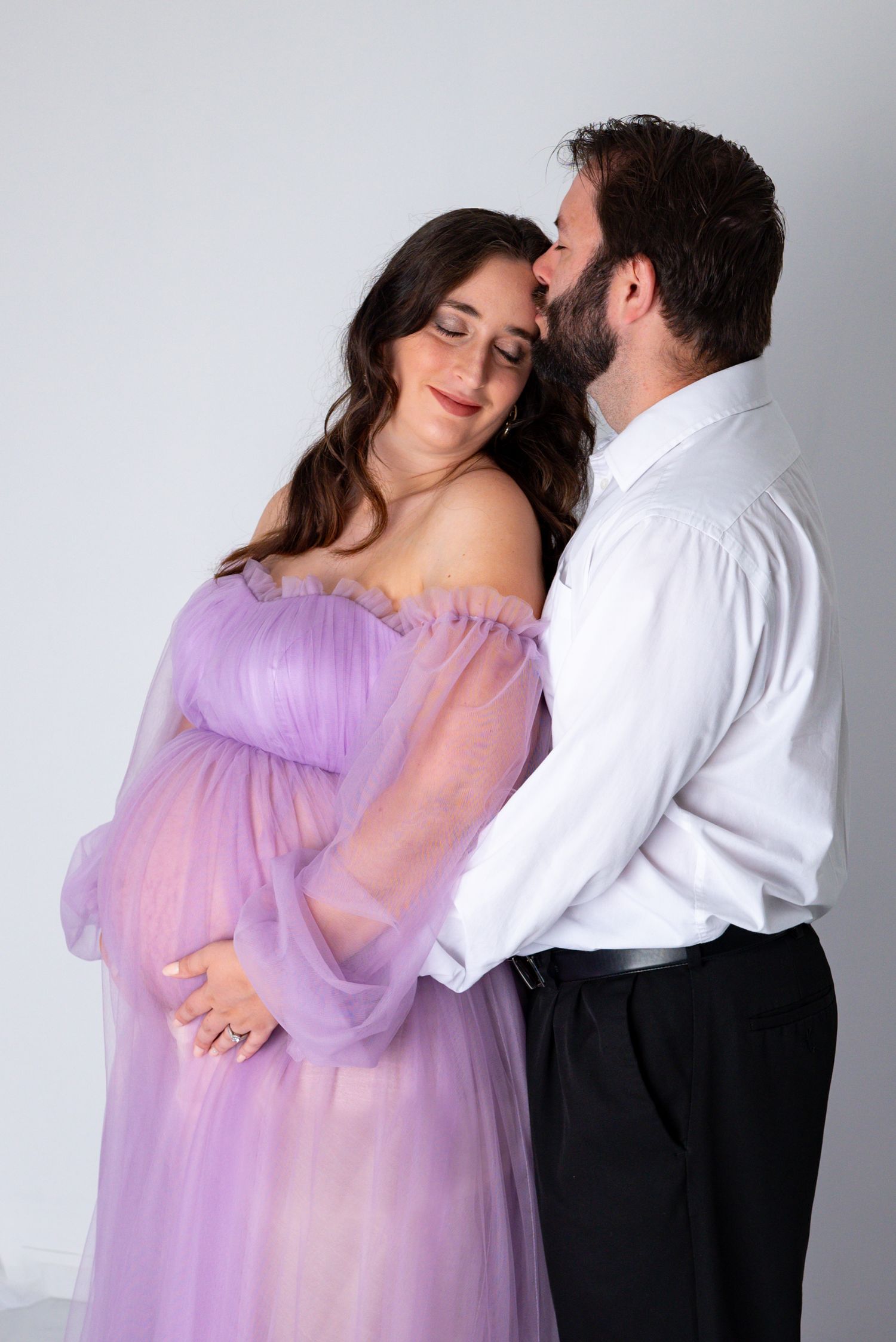A bearded man kisses his pregnant wife on the cheek as she smiles in a lavender off-shoulder tulle gown.