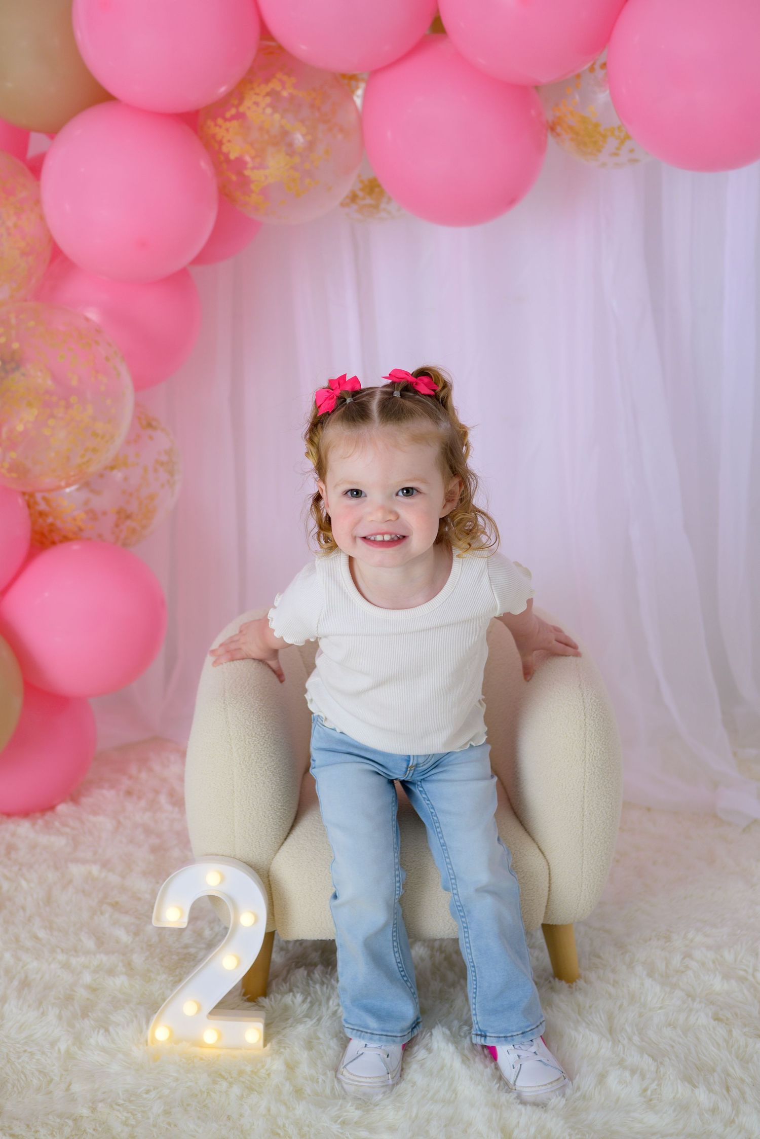 A smiling toddler girl sits in a cream chair beside a lit number 2, surrounded by pink and gold balloons for her 2nd birthday.