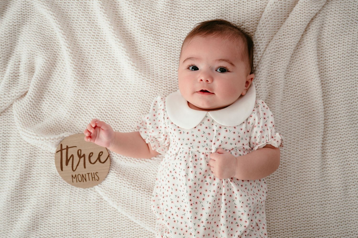 Baby girl in white floral dress holds a wooden three months milestone disc, lying on a cream knit blanket.