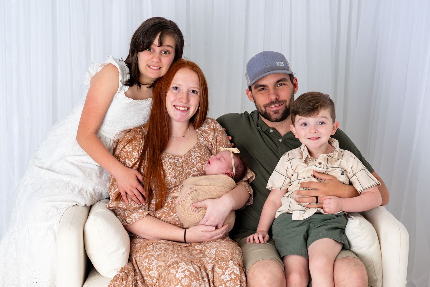 Family of five posing on white couch with newborn baby, featuring redhead mom, dad in green shirt, and two older children.