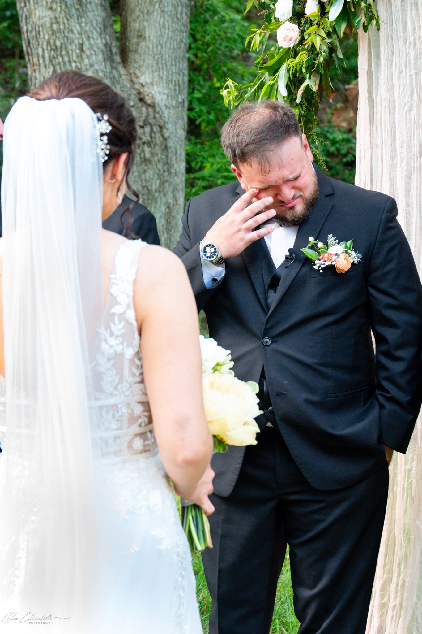 Emotional groom wipes tears at outdoor wedding ceremony while bride holds bouquet of flowers.
