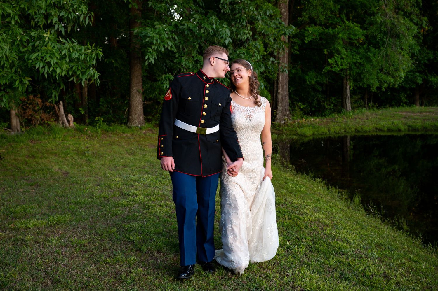 Marine in dress uniform holds hands with bride in white gown outdoors near pond.
