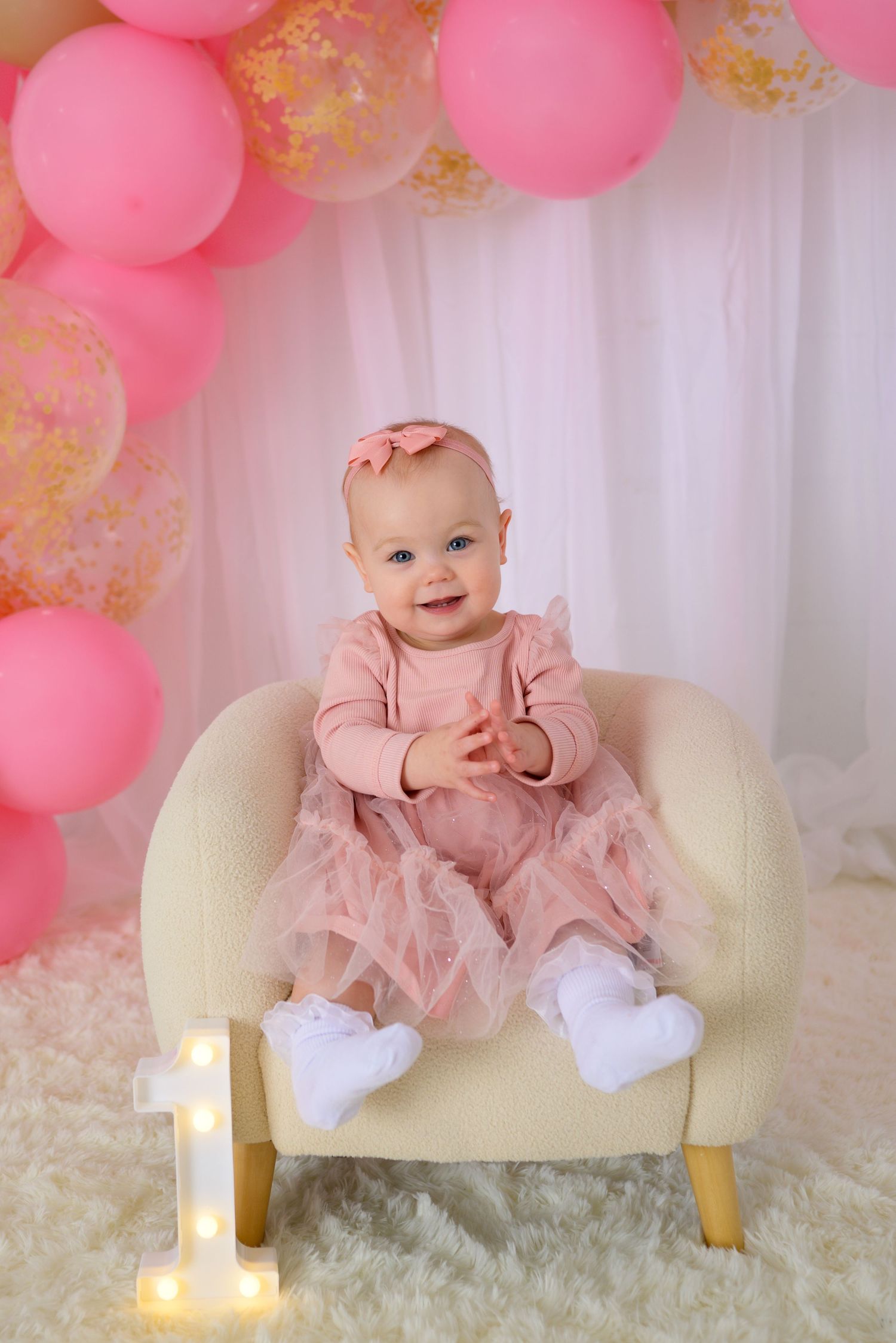 Happy baby girl in pink dress sitting in cream chair with lit number 1 and pink balloon arch.