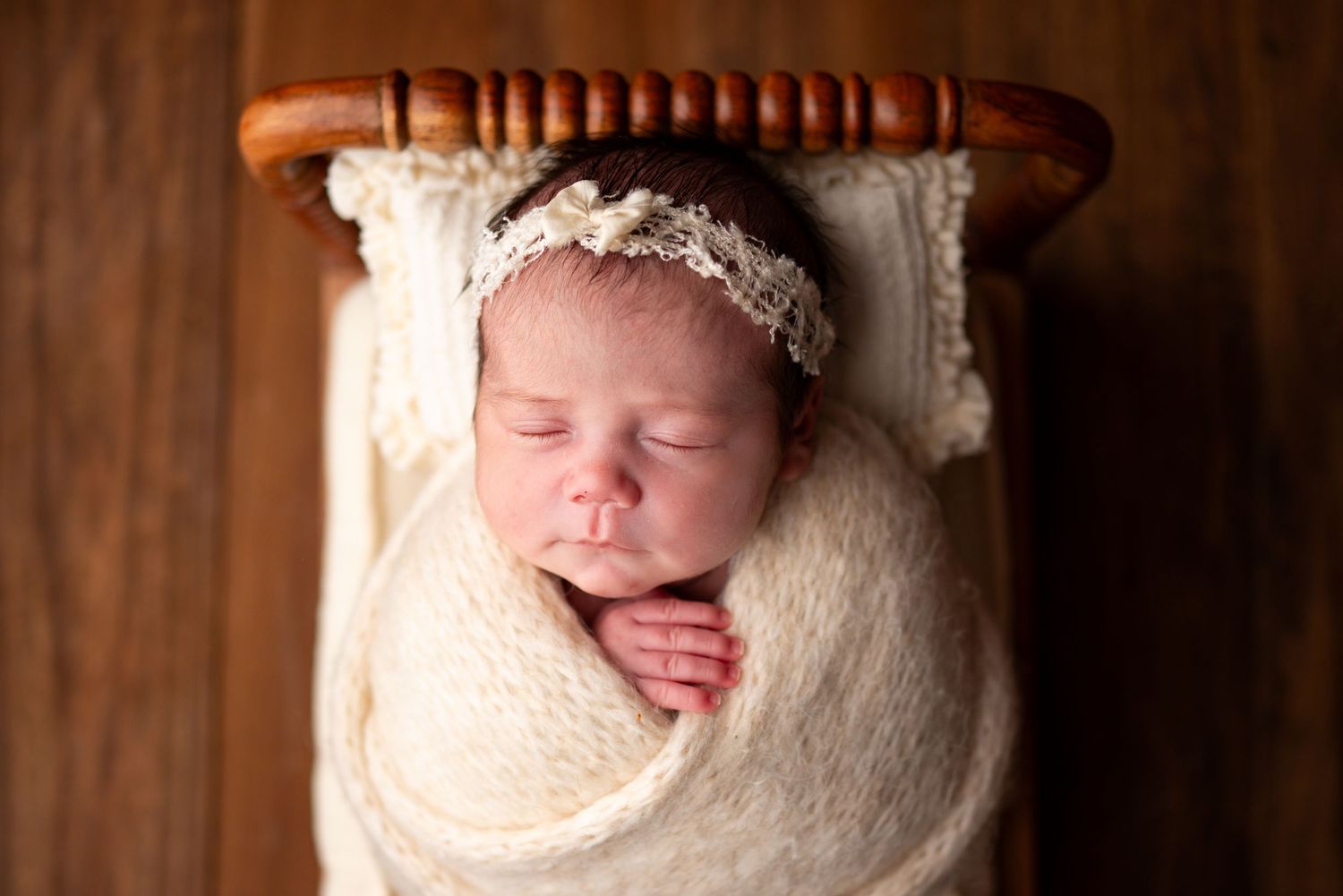 Newborn baby girl swaddled in cream wrap with lace headband, sleeping peacefully in a wooden mini bed.