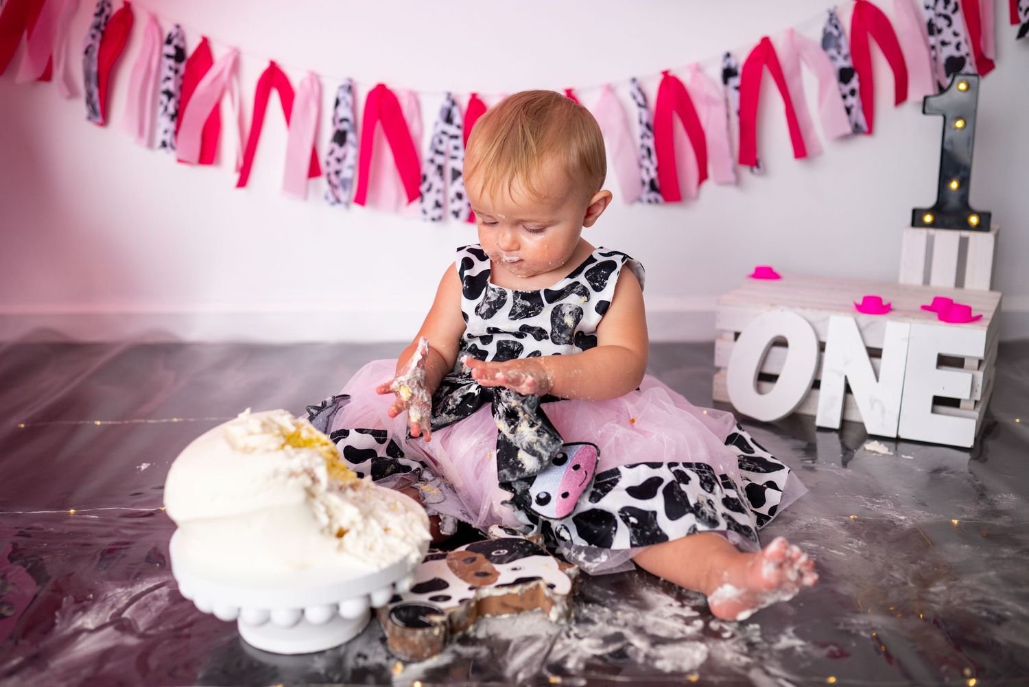 Young girl in floral dress celebrating first birthday with cake smash photoshoot and pink decorations.