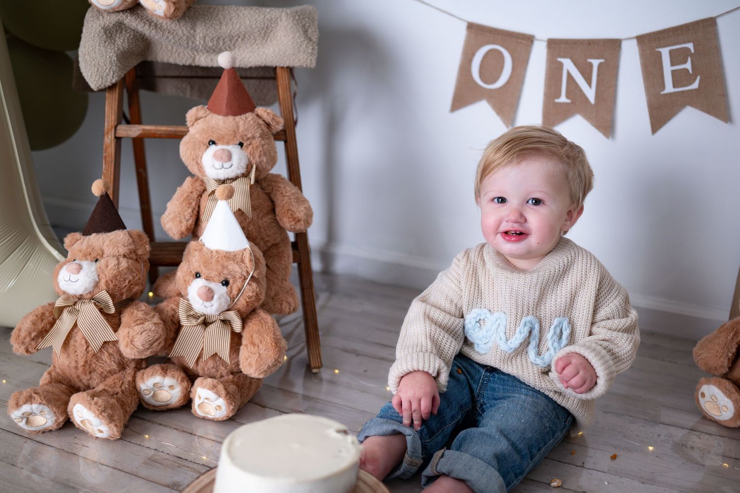Cheerful baby boy in ONE sweater sits beside teddy bears stacked on chair for first birthday photos.