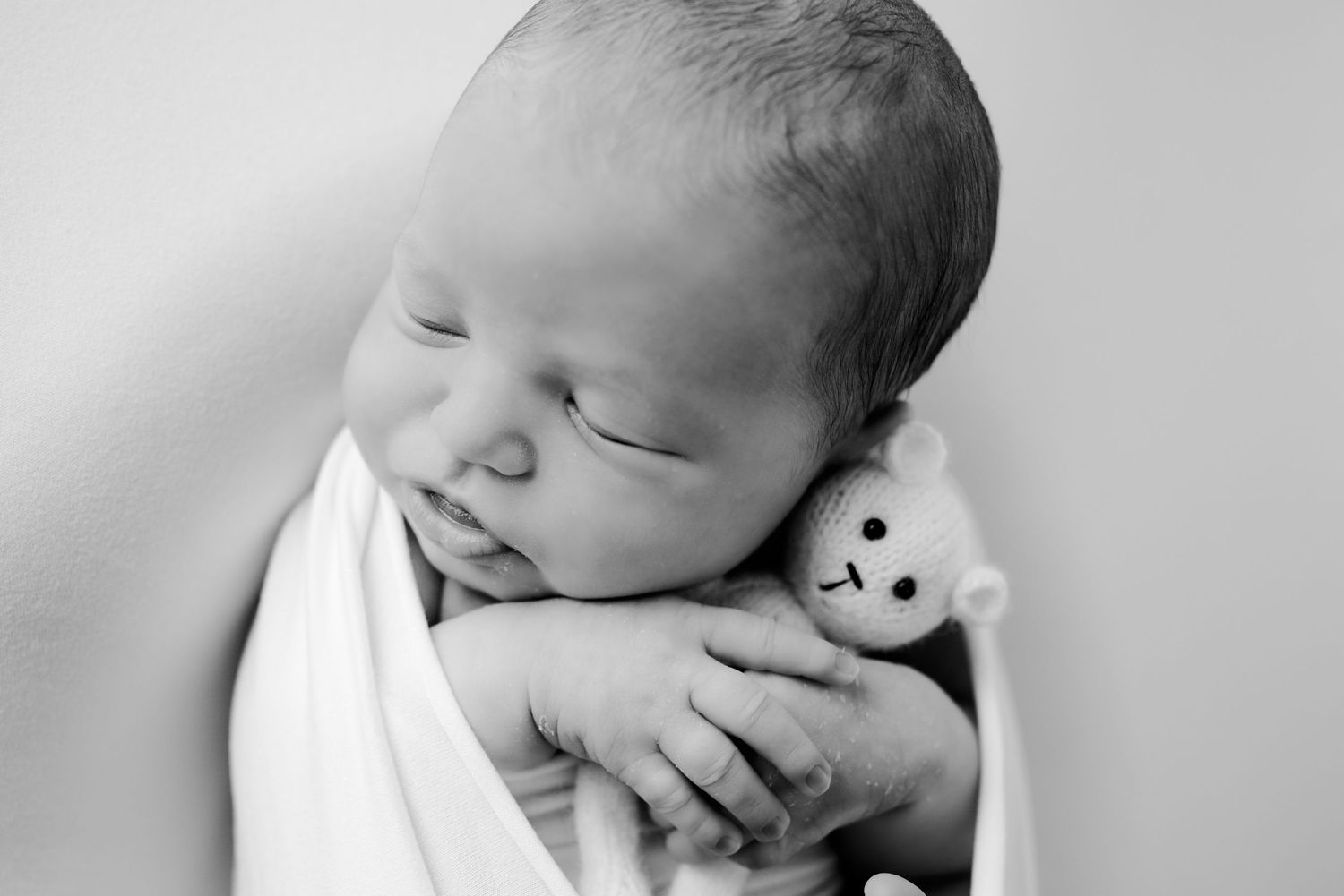 Black and white portrait of swaddled newborn baby sleeping while clutching a small knit bear.