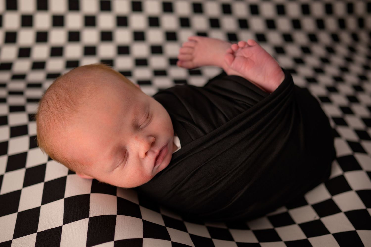 Sleeping newborn baby swaddled in black wrap on checkered black and white backdrop.