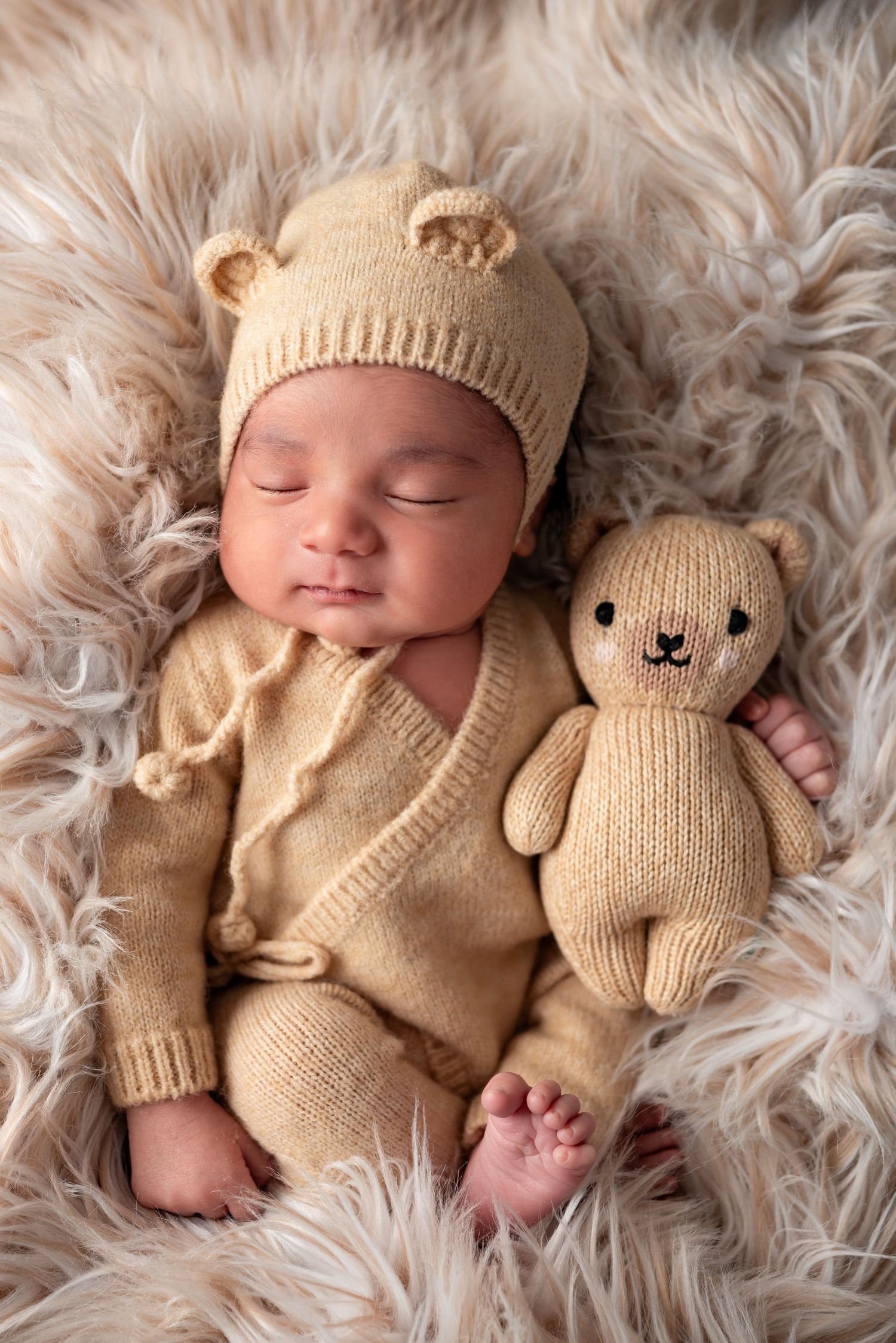 Sleeping newborn baby in beige bear ear hat and knit outfit lying beside teddy bear on fur.
