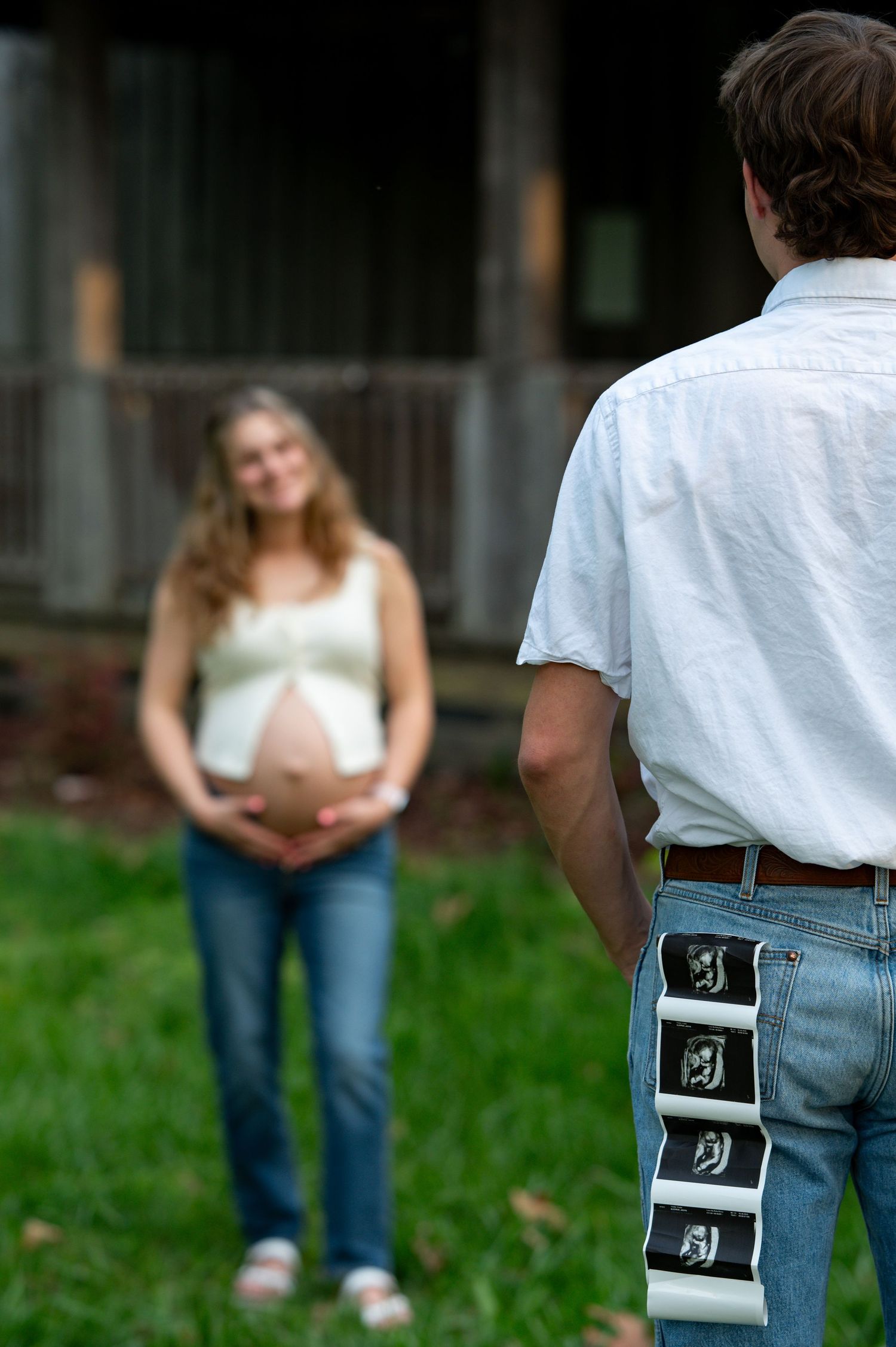 Pregnant woman stands smiling in yard as her partner faces her with ultrasound strip photos tucked in his pocket.