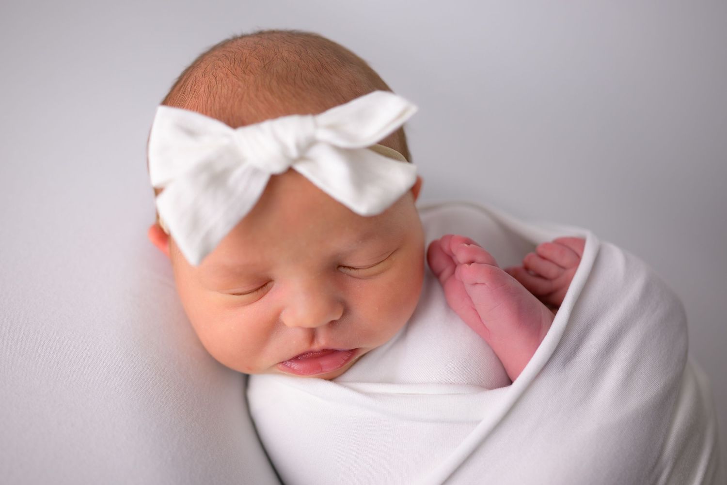 Sleeping newborn baby girl swaddled in white wrap with a white bow headband.