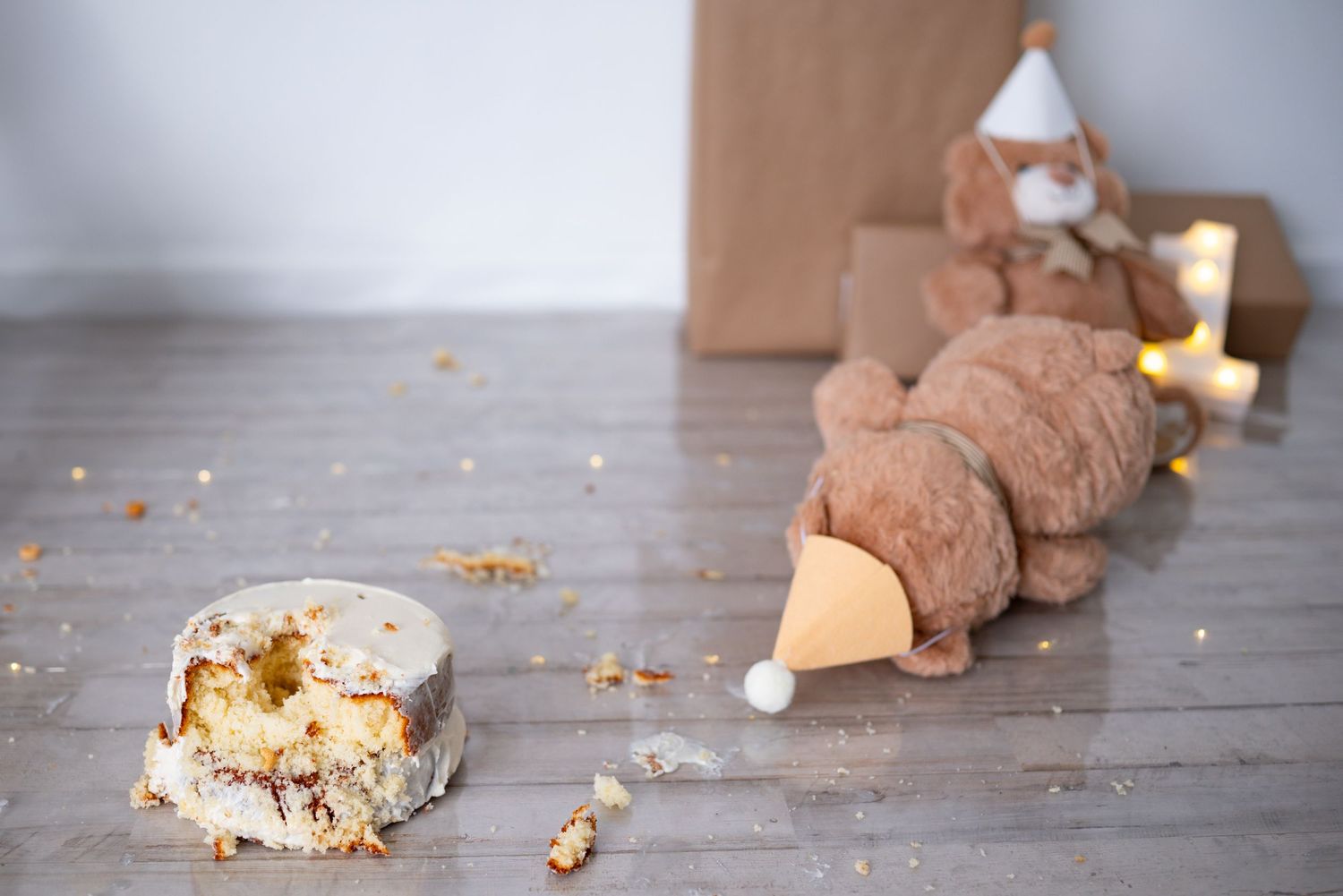 Smashed first birthday cake with crumbs scattered beside a teddy bear with party hat on wood floor.