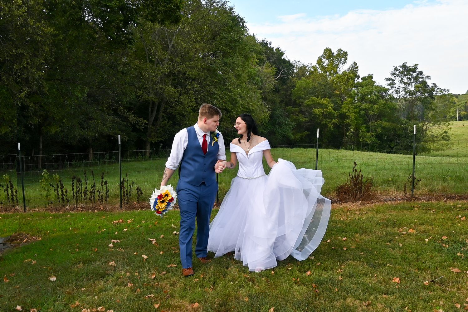 Bride and groom walking hand in hand on green lawn, bride in flowing white gown, groom in blue suit with red vest.