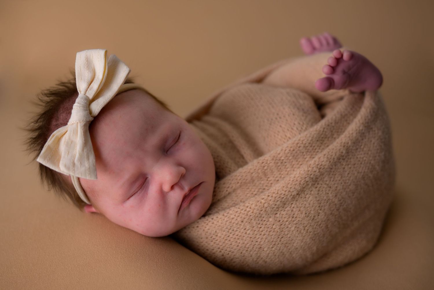 Sleeping newborn baby swaddled in tan wrap with white bow headband on brown background.