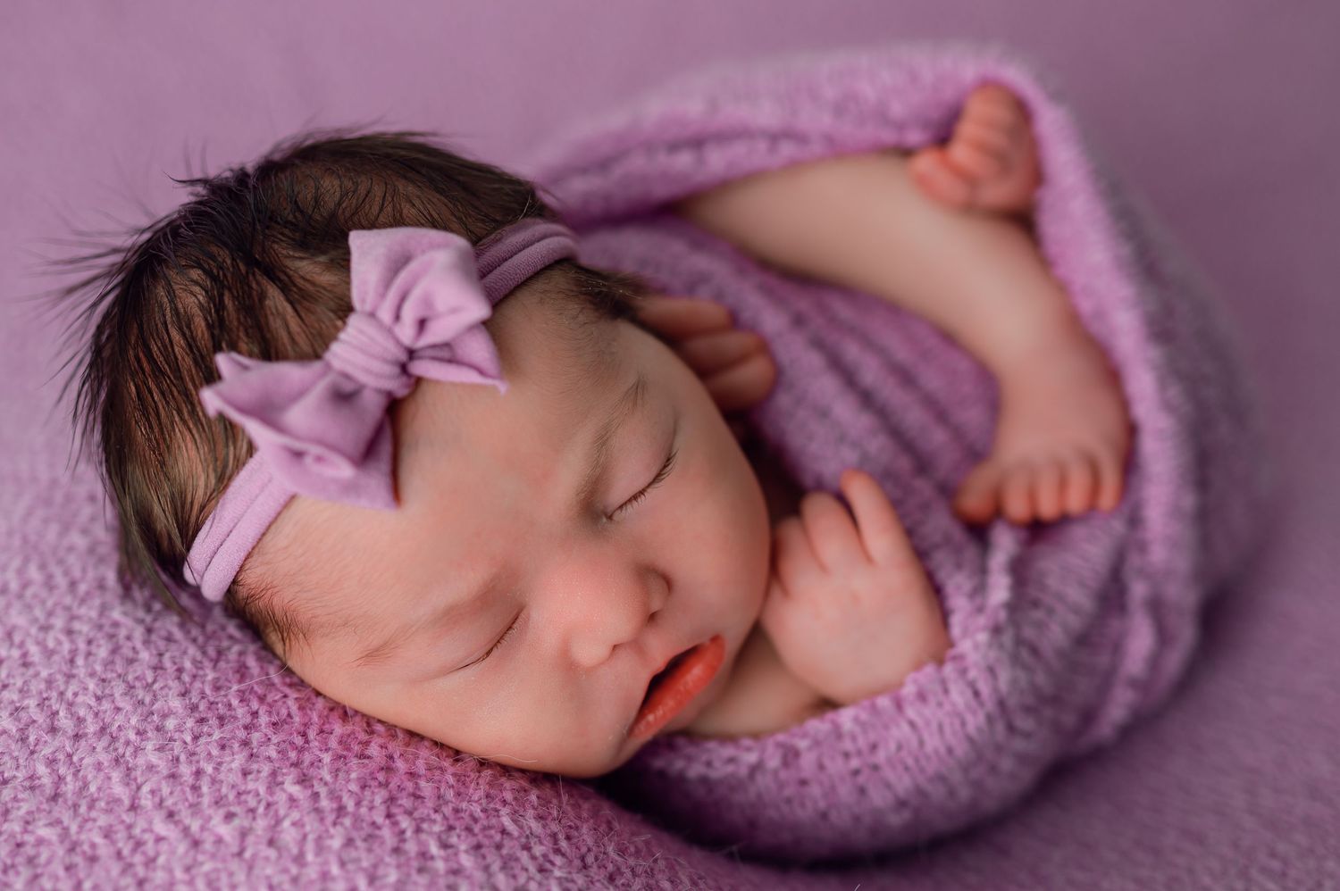 Sleeping newborn baby girl wrapped in purple swaddle with bow headband on lavender background.