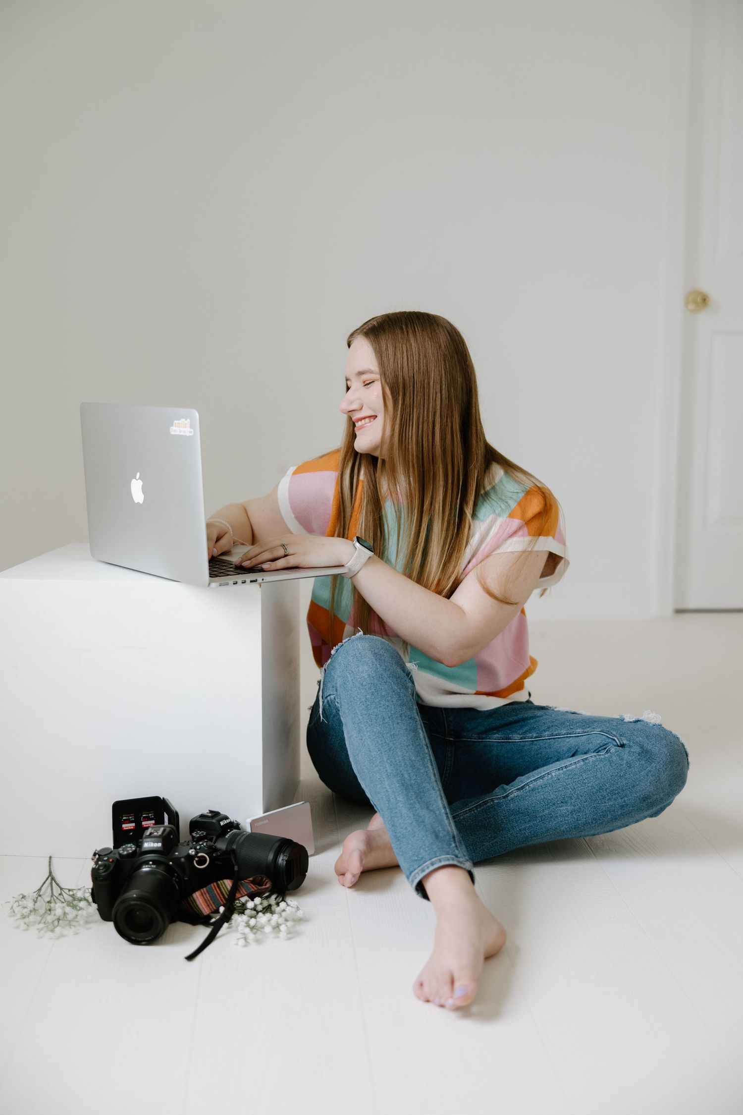 Young woman sitting cross-legged on floor, smiling at MacBook laptop, with DSLR camera nearby, wearing colorful striped shirt.