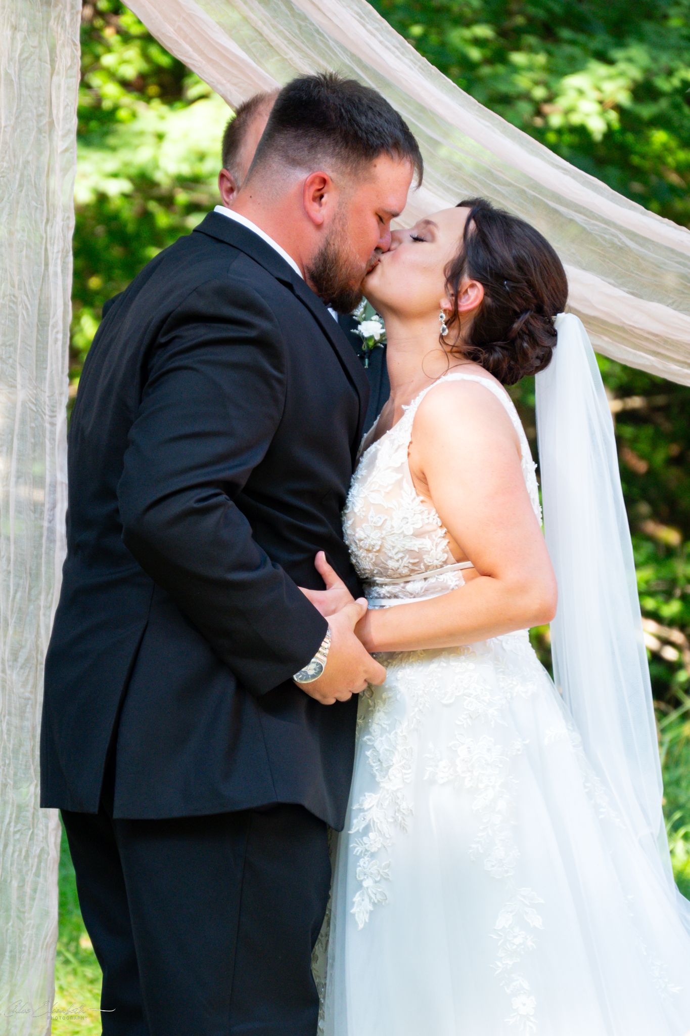Bride and groom share first kiss at outdoor wedding ceremony under draped fabric arch surrounded by lush greenery.