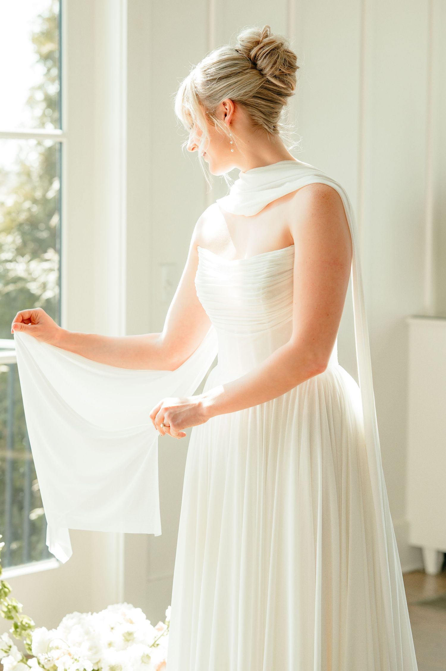 Bride in strapless white tulle gown holding veil, elegant updo hairstyle, standing by bright window on wedding day.