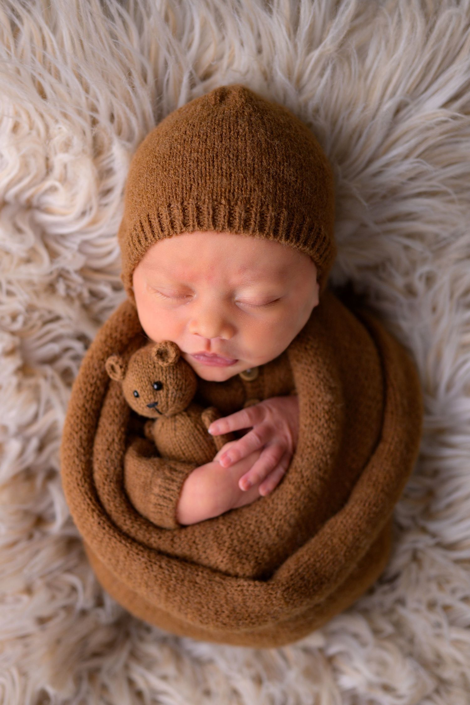 Newborn baby sleeping in brown knit wrap and beanie hat cuddling a small knitted teddy bear on fluffy rug.