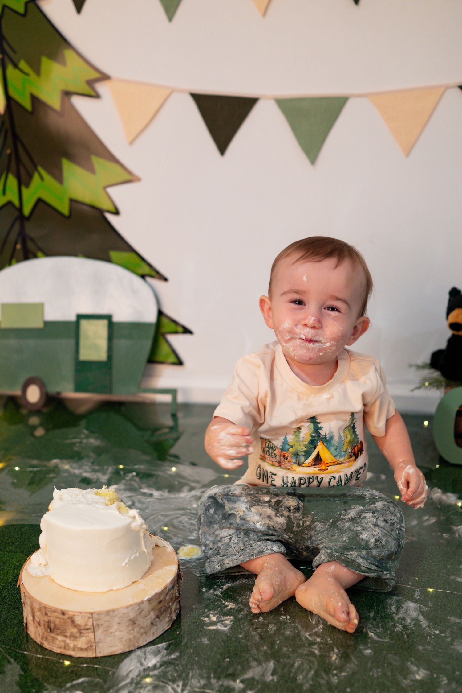 One-year-old boy with cake on his face sits beside his smash cake at a camping-themed birthday photoshoot.